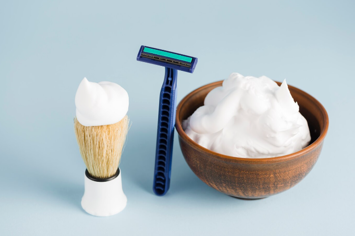 A close-up of shaving foam, a shaving brush, and a razor arranged neatly against a solid background.